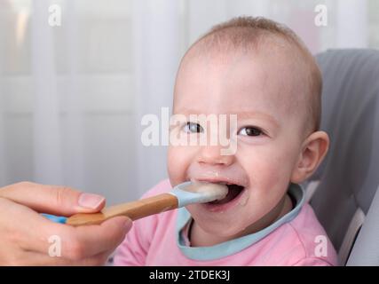 La mano della madre dà da mangiare al porridge di grano saraceno. Il bambino sorridente ride e guarda la telecamera. Spazio di copia per il testo Foto Stock