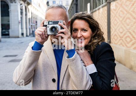 Una coppia di turisti adulti di medie dimensioni che scattano foto con una fotocamera vintage che guarda la fotocamera. Donna anziana che abbraccia un uomo mentre fa una fotografia in una strada cittadina Foto Stock