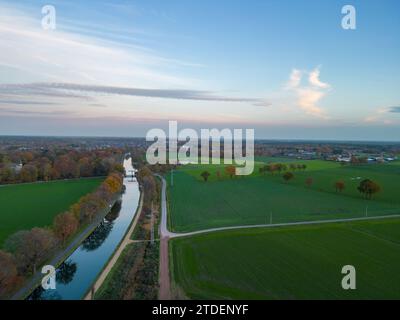 Questa immagine offre una vista aerea di un tranquillo canale che attraversa campi lussureggianti al crepuscolo. Il canale è delimitato da alberi ben rifilati, che riflettono la luce soffusa del cielo. I campi sono un verde intenso, indicativo di terreno ricco e fertile e di un'attenta coltivazione. Un piccolo percorso corre parallelo al canale, invitando lo spettatore a entrare nella scena. In lontananza, i deboli contorni delle turbine eoliche e delle strutture suggeriscono la presenza di una vicina città o zona industriale, mescolando paesaggi rurali e sviluppati. Crepuscolo sul canale e sui campi. Foto di alta qualità Foto Stock