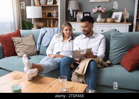 Giovani coppie entusiaste sedute sul divano indossando un tablet pc, celebrando la vittoria online, grandi offerte o il successo aziendale a casa. Coniuge Millennial Foto Stock