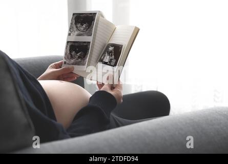 Una donna incinta è seduta su un divano vicino alla finestra nel soggiorno e guarda un'immagine ecografica del suo feto Foto Stock