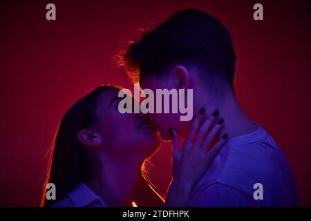 Adorabile giovane uomo e donna che si baciano con tenerezza, mostrando amore e cura su sfondo rosso con luce al neon Foto Stock