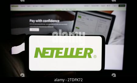 Persona che possiede il cellulare con il logo della società Neteller, una piattaforma di pagamenti globale, davanti alla pagina Web aziendale. Concentrarsi sul display del telefono. Foto Stock