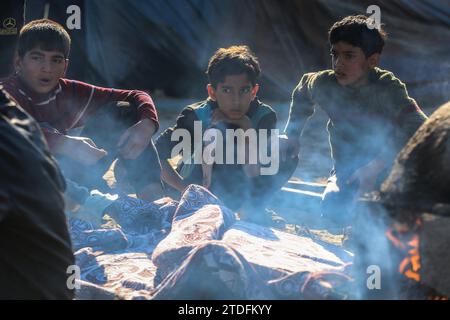 Rafah, territori palestinesi. 18 dicembre 2023. I bambini guardano mentre le persone si radunano intorno ai forni di argilla per riservarsi il loro turno di preparare cibo e cuocere pane, in condizioni di energia elettrica, acqua e rifornimenti alimentari molto limitate, in un campo per palestinesi sfollati a Rafah, dove la maggior parte dei civili si è rifugiata dal bombardamento israeliano in corso. Credito: Mohammed Talatene/dpa/Alamy Live News Foto Stock