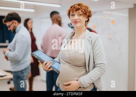 Donna d'affari incinta che tiene un portatile in posa in ufficio. Persone che collaborano in un ambiente di ufficio innovativo sul retro. Foto Stock