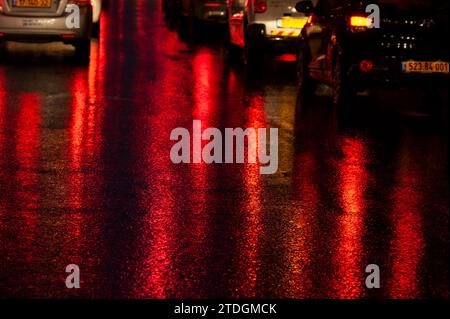 Strada asfaltata bagnata e astratta, colorata di notte da luci di posizione posteriori rosse e semafori. Foto Stock