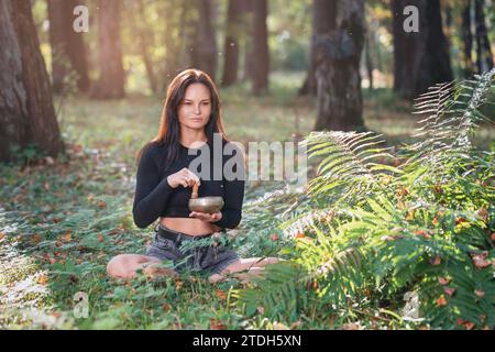 Una donna fa una meditazione sana con una ciotola tibetana, allevia lo stress e migliora lo stato generale del corpo, pratica yoga nel parco su una W soleggiata Foto Stock