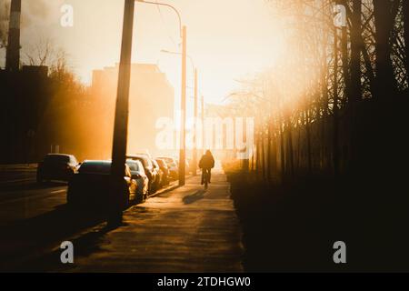 Un paesaggio urbano illuminato dal luminoso sole mattutino, dove un uomo solitario cavalca una bicicletta per lavorare in una mattinata nebbiosa. Stile di vita. Un'ombra. Foto Stock