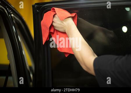 Pulizia auto. Rimozione della polvere dal vetro della macchina. Uno straccio rosso in mano. Lavoro di lavaggio auto. Foto Stock