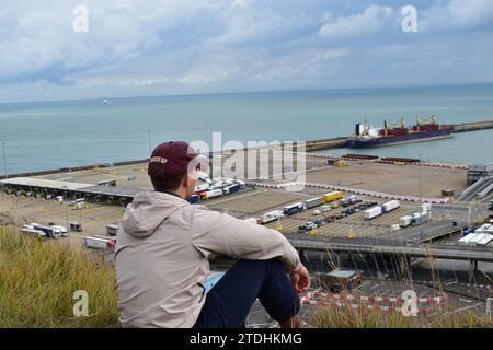 Un giovane uomo che indossa un berretto bordeaux, una giacca da pioggia beige chiaro e pantaloni blu scuro che guarda il porto di dover dalla scogliera Foto Stock