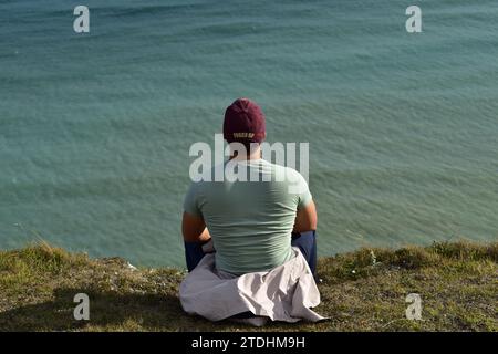 Giovane uomo che indossa pantaloni blu scuro, una t shirt verde chiaro e un berretto bordeaux sul bordo delle bianche scogliere di dover affacciato sul mare Foto Stock