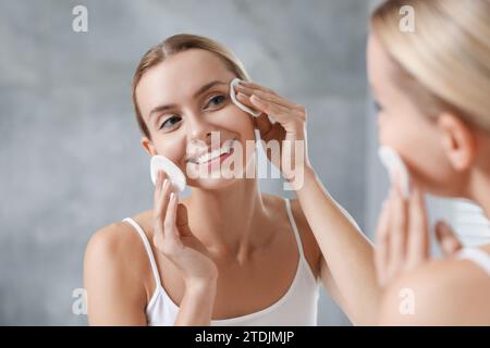 Donna sorridente che si toglie il trucco con tamponi di cotone davanti allo specchio in interni Foto Stock