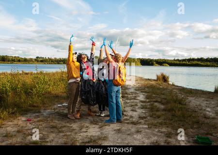 Questa immagine edificante ritrae un gruppo di quattro volontari, due donne e due uomini, che celebrano la loro pulizia di successo in un lago. Sono diversi per età ed etnia, tra cui un giovane uomo caucasico, una donna ispanica e un uomo caucasico anziano. Ognuno alza le mani con i guanti in trionfo, davanti allo sfondo panoramico di un lago azzurro e di un cielo parzialmente nuvoloso. Il loro abbigliamento informale e i sacchetti pieni di rifiuti raccolti parlano del loro duro lavoro e del loro impegno nel preservare l'ambiente naturale. Vittoria per l'ambiente: Successo di pulizia del gruppo. Foto di alta qualità Foto Stock