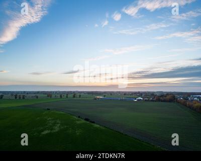 Questa immagine serena mostra la vasta distesa di un paesaggio rurale al tramonto. Il cielo, dipinto con sfumature delicate di arancio e blu, fornisce uno sfondo drammatico ai tranquilli campi sottostanti. Una lontana linea di alberi e un piccolo gruppo di edifici sulla destra introducono un elemento umano nella scena, suggerendo una piccola comunità o una fattoria. I campi aperti sono segnati dalle lunghe ombre del sole che tramonta, esaltando la consistenza della terra e sottolineando la pacifica fine giornata. Tramonto su un paesaggio rurale. Foto di alta qualità Foto Stock