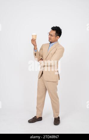 Un uomo d'affari asiatico millenario di successo e intelligente è una tuta da lavoro che sta in piedi con una tazza di caffè in mano contro una schiena bianca isolata dello studio Foto Stock