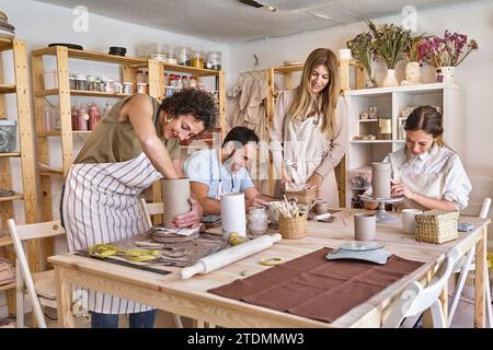 Gruppo entusiasta di quattro argilla modellante in uno studio di ceramica ben illuminato pieno di scaffali e strumenti. Foto Stock