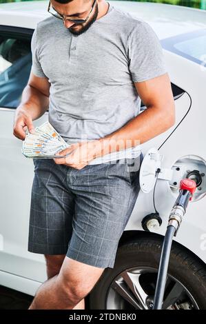 Conducente preoccupato a contare quanti soldi deve spendere per il carburante. I prezzi aumentano a causa della mancanza di carburante. Uomo triste in abiti casual e occhiali da sole che contano dollari nelle sue mani. Foto Stock