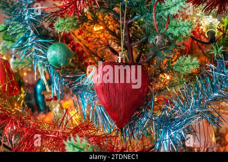 Primo piano di un albero di Natale completamente decorato con un prominente bauble rosso a forma di cuore tra decorazioni e luci multicolore, bokeh di sfondo. Foto Stock