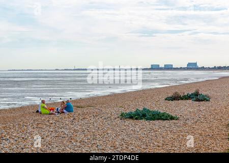 Gruppo di persone che si rilassano sulla spiaggia di Littlestone nel Kent, Inghilterra sud-orientale, Regno Unito, con la centrale nucleare di Dungeness visibile all'orizzonte. Foto Stock