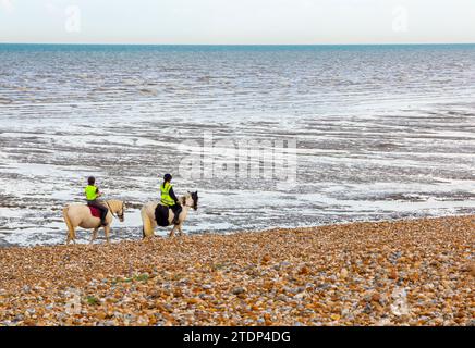 Due donne cavalcano a cavallo su una spiaggia di ciottoli con un mare calmo sullo sfondo. Foto Stock