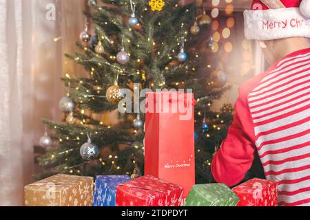 La vista dei ragazzi con il cappello di Babbo Natale apre un regalo di fronte all'albero di Natale illuminato, nella calda atmosfera di Natale Foto Stock