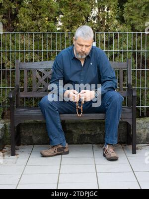 Uomo con capelli grigi e barba piena, 50-59 anni, seduto su una panchina sulla terrazza esterna e pregando il Rosario Foto Stock