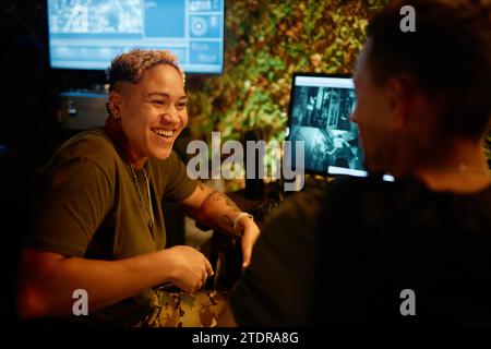 Felice giovane donna militare che guarda un collega maschio durante la discussione mentre entrambi sono seduti davanti agli schermi del computer nella sala di sorveglianza Foto Stock