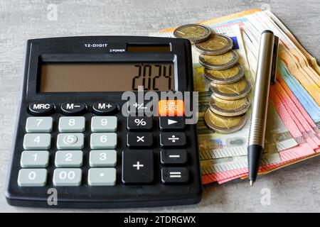 Calcolatore con il numero 2024 in esposizione, penna, banconote in euro e monete su sfondo grigio Foto Stock