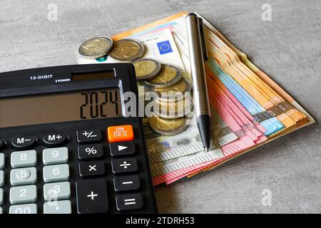 Calcolatore con il numero 2024 in esposizione, penna, banconote in euro e monete su sfondo grigio Foto Stock