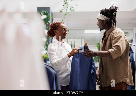 Uomo afro-americano che riceve assistenza da un lavoratore di abbigliamento mentre seleziona la giacca. Cliente del centro commerciale che chiede aiuto ad un assistente per scegliere un nuovo outfit in boutique Foto Stock