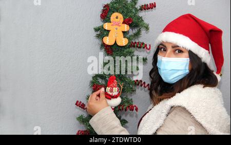 Donna latina con maschera protettiva, maglione e cappello di natale su sfondo grigio con decorazione natalizia, nuovo normale Foto Stock