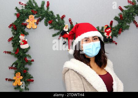 Donna latina con maschera protettiva, maglione e cappello di natale su sfondo grigio con decorazione natalizia, nuovo normale Foto Stock