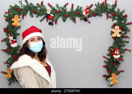 Donna latina con maschera protettiva, maglione e cappello di natale su sfondo grigio con decorazione natalizia, nuovo normale Foto Stock