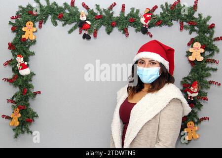 Donna latina con maschera protettiva, maglione e cappello di natale su sfondo grigio con decorazione natalizia, nuovo normale Foto Stock