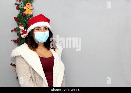 Donna latina con maschera protettiva, maglione e cappello di natale su sfondo grigio con decorazione natalizia, nuovo normale Foto Stock