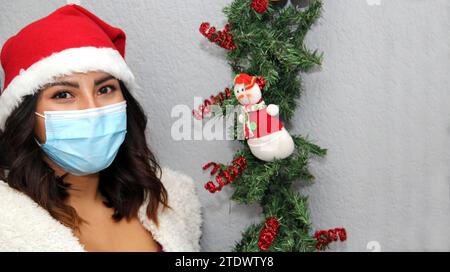Donna latina con maschera protettiva, maglione e cappello di natale su sfondo grigio con decorazione natalizia, nuovo normale Foto Stock