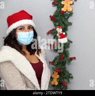 Donna latina con maschera protettiva, maglione e cappello di natale su sfondo grigio con decorazione natalizia, nuovo normale Foto Stock