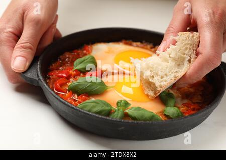 Donna che immerge un pezzo di pane nel delizioso Shakshuka al tavolo bianco, primo piano Foto Stock