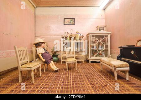 Un bambino seduto su una sedia in una sala musica con un pianoforte in una vecchia casa di bambole di legno tradizionale e antica. All'interno del Phra Tamnak Daeng, la Casa Rossa. Foto Stock