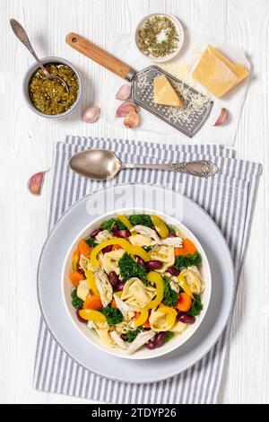 minestrone di pollo con tortellini e fagioli rossi, cavolo e peperone giallo in ciotola su tavolo di legno bianco con cucchiaio, pesto e parmesa Foto Stock