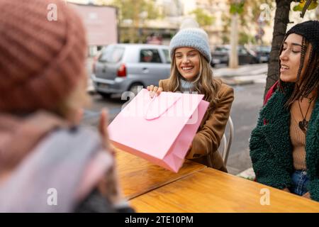Tre donne condividono la risata davanti a un drink caldo in un caffè all'aperto, mostrando uno stile di vita urbano rilassato. Foto Stock