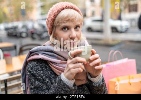 Una giovane donna bionda si gode una calda tazza di caffè in una strada cittadina, incarnando una rilassante giornata invernale. Foto Stock