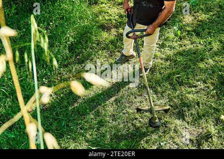 Giardiniere che taglia le erbacce in giardino con il rifinitore per erba con corda. Uomo che usa il rasaerba in giardino, tagliando erba verde. Foto Stock