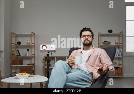 Un uomo bello, dai capelli scuri e di mezza età con abiti informali sta guardando un film usando il proiettore. Il tizio barbuto con gli occhiali guarda lo schermo del sistema Home Theater Foto Stock
