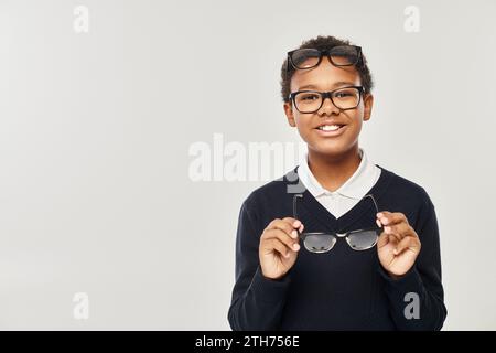 ragazzo afro-americano ottimista con maglione e occhiali che regge gli occhiali e guarda la fotocamera sul grigio Foto Stock