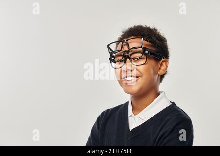 ragazzo afro-americano ottimista con maglione e occhiali che regge gli occhiali e guarda la fotocamera sul grigio Foto Stock