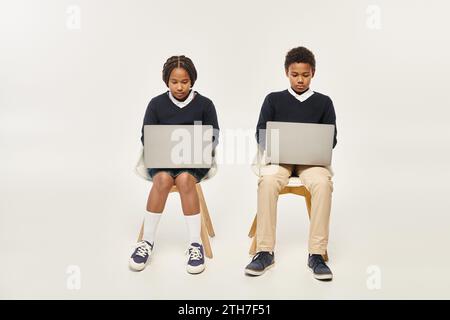 scolari afroamericani concentrati in uniforme che usano computer portatili e sono seduti su sfondo grigio Foto Stock