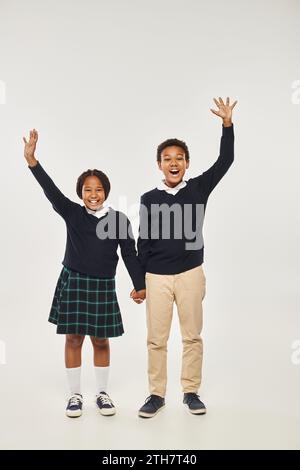 eccitati bambini afroamericani preadolescenti in uniforme che si tengono per mano su sfondo grigio Foto Stock