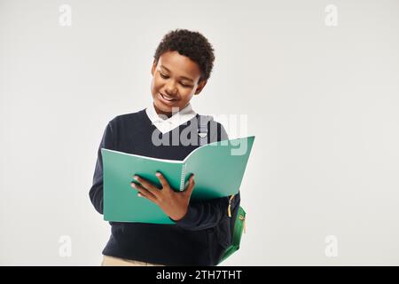 bravo scolaro afroamericano in uniforme che guarda un notebook e si trova su sfondo grigio Foto Stock