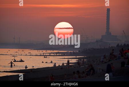 La spiaggia di Hove guarda ad ovest verso Shoreham, con il sole che tramonta sopra l'orizzonte e la gente che delizia la fine di una calda giornata estiva. Brighton e Hove. East Sussex, Inghilterra. Shoreham Power Station in lontananza Foto Stock